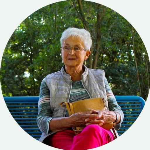 Foto: Ältere Dame sitzt fröhlich auf einer Parkbank bei schönem Wetter.