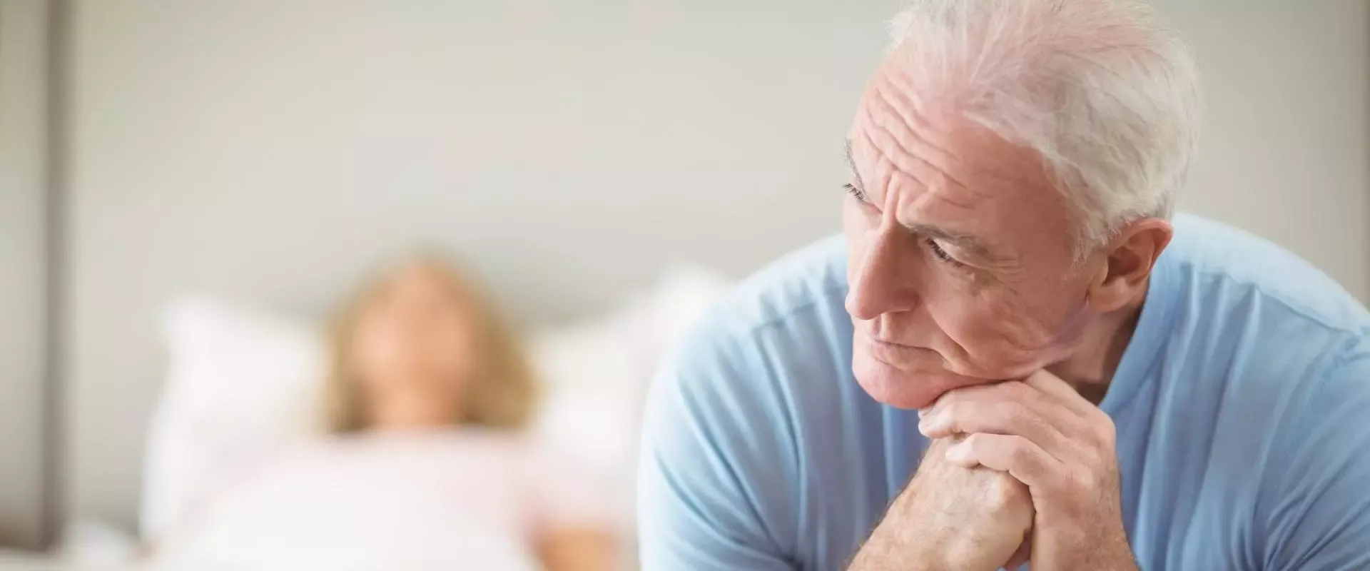 Mann sitzt grübelnd an der Bettkante, im Hintergrund liegt eine Frau im Bett