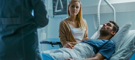 Mann liegt im Krankenhausbett, Partnerin sitzt daneben